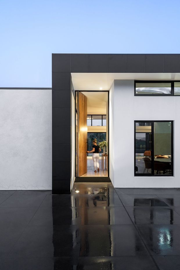 Contemporary residential facade photographed from outside, featuring a dark charcoal tiled or panelled entry surround, crisp white rendered walls, a tall timber pivot door, black-framed glazing, and a glossy dark tiled forecourt reflecting the architecture. Interior spaces are visible through the open entry and window, with warm timber finishes and furnishings. The image is taken after rain under a clear blue sky.