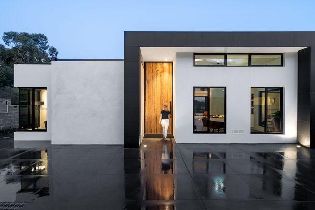 Contemporary single-storey residential facade with clean white rendered walls, dark charcoal framing and cladding, extensive black-framed glazing, and a warm timber entry door. The building opens onto a broad glossy dark paved terrace or forecourt, reflecting the facade and evening lighting. Minimal landscaping and a flat-roofed, cubic composition emphasize the modern design.