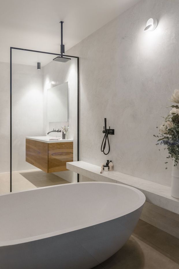 A refined contemporary bathroom featuring a large freestanding oval bathtub in the foreground, a walk-in shower enclosed by black-framed glass, a floating timber vanity with integrated basin, wall-mounted black shower fixtures, textured light plaster walls, and warm neutral flooring. Minimal decor and soft lighting create a calm, luxurious atmosphere.