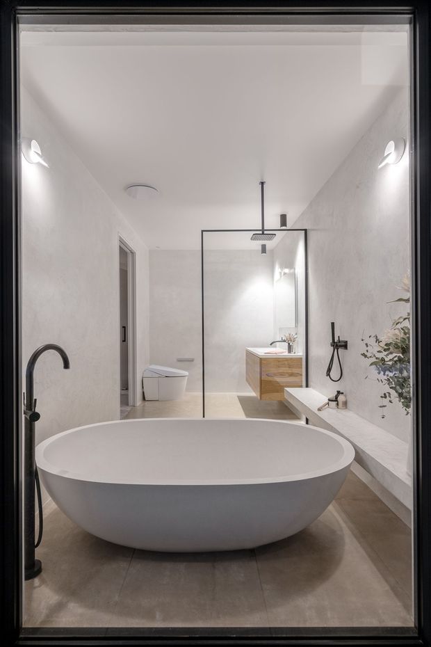 A refined contemporary bathroom viewed through a dark-framed glazed opening. The room features a large freestanding oval bathtub in the foreground, a walk-in shower with a frameless glass screen and ceiling-mounted showerhead, a wall-mounted timber vanity with integrated basin, wall-hung toilet, and a long built-in ledge with dark tapware and decorative greenery. The palette is predominantly white, light grey, warm timber, and charcoal accents, with smooth plastered walls and large-format stone-look flooring.