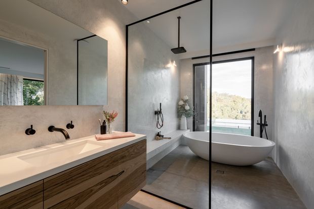 Contemporary bathroom with a double vanity, wall-mounted black fixtures, large mirror, frameless glass shower enclosure, freestanding oval bathtub, textured plaster walls, and warm wood-grain cabinetry. A large window provides an outdoor view and natural light.