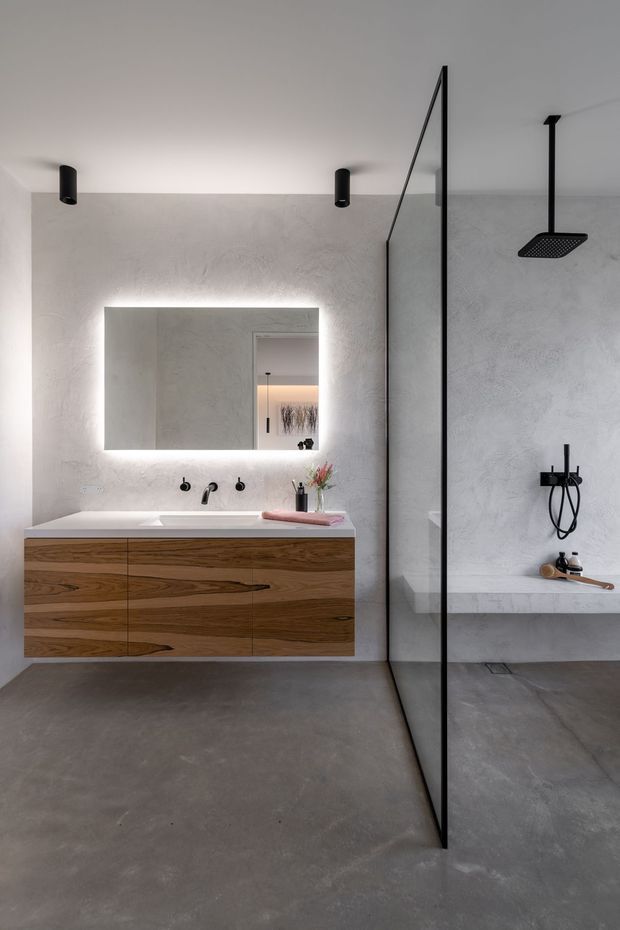 A contemporary minimalist bathroom with a floating timber vanity, integrated white basin, black wall-mounted tapware, illuminated frameless mirror, and a large walk-in shower enclosed by a slim black-framed glass screen. The room features smooth grey concrete-look flooring and textured pale grey wall finishes, with recessed and cylindrical black lighting.