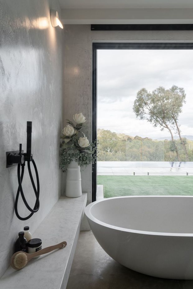 A refined contemporary bathroom featuring a freestanding oval bathtub beside full-height glazing, a wall-mounted black bath/shower fitting, and a long pale stone ledge with bath accessories and a white vase of flowers. Textured light grey walls, dark flooring, and expansive views toward landscaped grounds create a calm, spacious atmosphere.