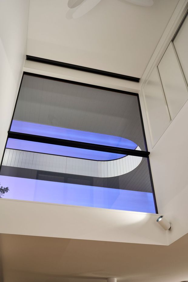 Upward-looking interior view of a bright double-height space with a large glazed opening or skylight, black-framed glazing, and a curved architectural overhang visible beyond. White walls and ceiling surfaces are accented by recessed and directional lighting.