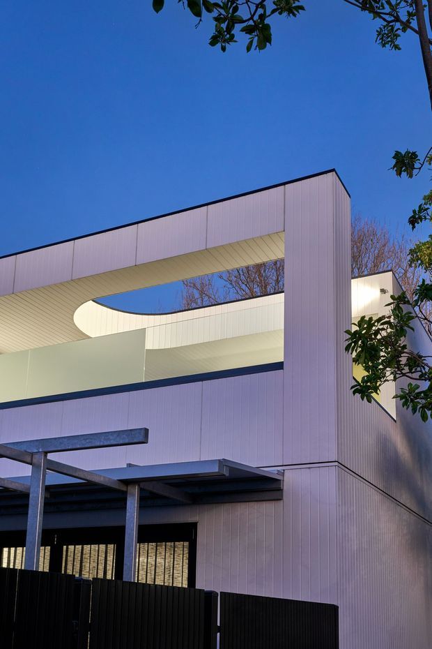 A contemporary multi-level building exterior photographed at dusk, featuring pale vertical panel cladding, dark metal framing and fencing, deep roof overhangs, and a distinctive curved open-air void cut through the upper structure. Warm interior lighting is visible through openings, with mature trees framing the facade.