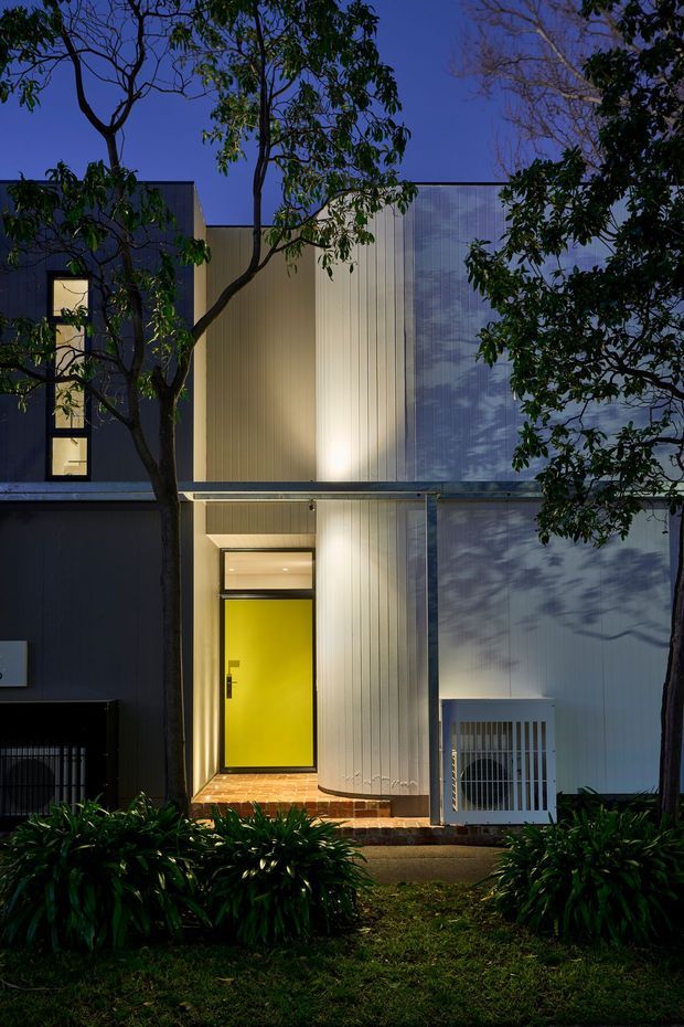 A dusk photograph of a contemporary multi-storey residential facade, framed by mature trees and garden planting. The building features dark cladding, pale vertically ribbed metal or composite wall panels, large narrow windows, a projecting horizontal canopy, and a striking bright yellow entrance door set above a brick-paved threshold.