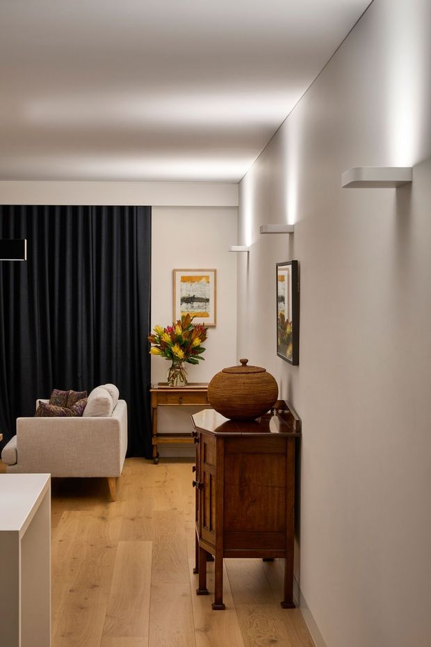 A refined living room or lounge area viewed along a narrow circulation zone, with pale timber flooring, warm off-white walls and ceiling, full-height black curtains, a light upholstered armchair, and dark traditional timber console furniture. Integrated linear wall lighting and small floating shelves create a softly illuminated contemporary backdrop, while framed artwork, flowers, and a large ribbed ceramic vessel add warmth and character.