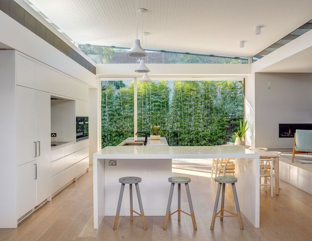 Bright open-plan kitchen and living area centred on a large white island with integrated sink and three timber-and-metal stools. The kitchen features extensive flush white cabinetry, pale timber flooring, a light benchtop, and minimalist pendant lights. Full-height glazing and clerestory windows open the room to a dense green garden, while a partially visible living zone with built-in seating and a fireplace sits to the right.