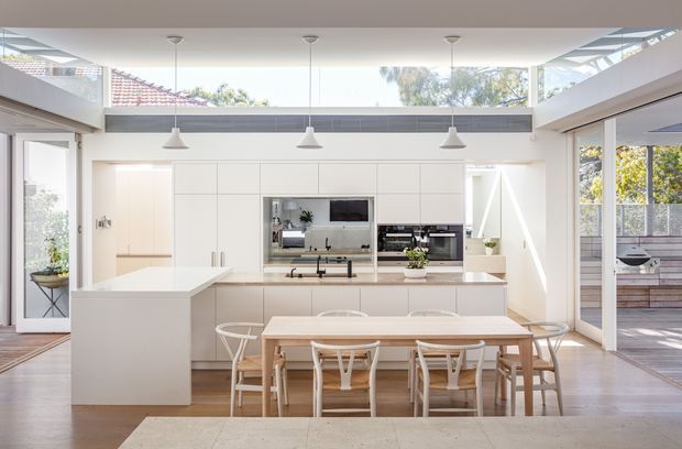 Bright open-plan kitchen and dining area with white built-in cabinetry, a large island, integrated appliances, timber dining table and light timber chairs. The space features extensive glazing, a high ceiling with clerestory windows, three pendant lights, and a covered outdoor area visible beyond the sliding doors.