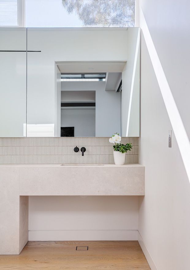 Bright minimalist bathroom vanity area with a large frameless mirror, pale stone or concrete benchtop, integrated wall-mounted basin, narrow vertical mosaic tile splashback, matte black tapware, white walls, and light timber flooring. A small potted orchid sits on the vanity. The mirror reflects a high, open interior with skylight-like glazing.