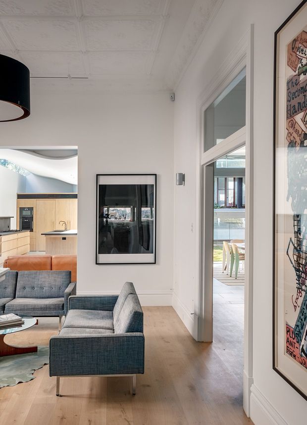 A bright, refined open-plan living area in a heritage-style home, viewed toward a compact kitchen and an adjoining outdoor deck. The room features white walls, ornate pressed-tin ceiling panels and decorative cornices, pale timber flooring, a blue-grey upholstered armchair and sofa, a tan leather sofa, framed artwork, and a black pendant light. Glazed internal and external openings bring in natural light and provide views to the deck and garden.