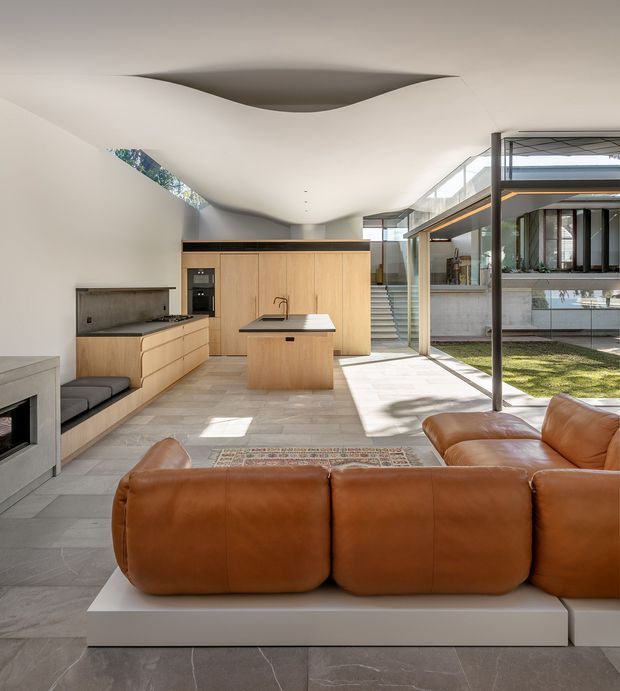 Bright open-plan kitchen and living area with a warm timber kitchen, central island, integrated appliances, and a large caramel leather sectional sofa in the foreground. The space has a sculptural curved ceiling, expansive glazing, pale stone-look flooring, and views toward a landscaped courtyard and adjacent modern structure. A fireplace and built-in bench seating add to the living zone.