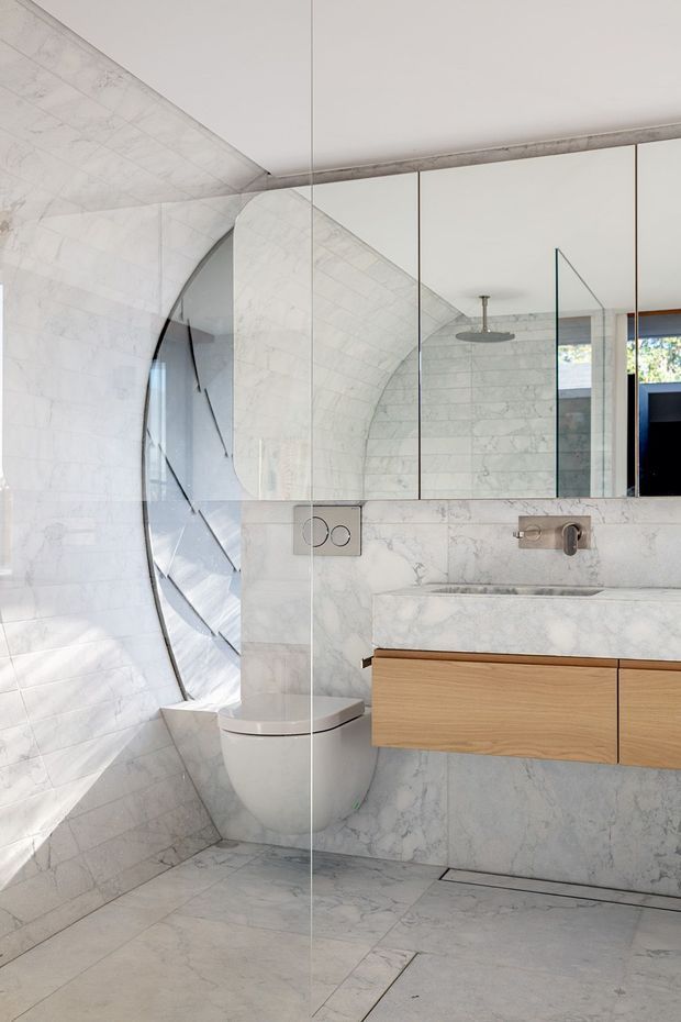 A contemporary bathroom with dramatic curved white marble-clad walls and ceiling, creating a sculptural arched enclosure around a wall-hung toilet. A floating timber vanity with a pale stone integrated basin sits beneath a large mirrored wall, with brushed metal wall-mounted tapware and a concealed cistern. The space is bright, refined, and predominantly monochromatic with warm timber detailing.