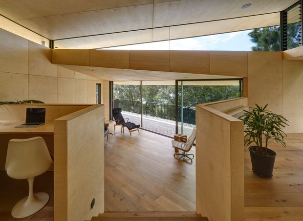 Warm, minimalist timber-lined interior with a compact workspace in the foreground and a sunken living area beyond. Pale timber wall and ceiling panels, built-in cabinetry and partitions, expansive glazing, and a leafy outlook create an airy, natural atmosphere. A desk with laptop, sculptural chair, lounge chairs, and indoor plants are visible.