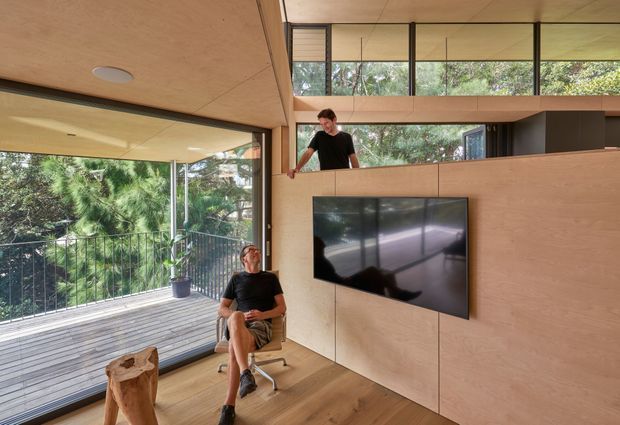 A warm, timber-lined contemporary interior featuring a compact living or media area opening onto a glazed balcony. Plywood wall and ceiling panels, integrated cabinetry, a wall-mounted television, and timber flooring create a cohesive built-in fit-out. A mezzanine-like upper level with clerestory glazing overlooks the room, while abundant glazing brings in views of surrounding trees. Two people are present, one seated in an office chair and another leaning over the upper platform.