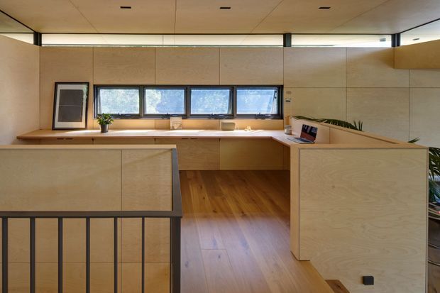 A warm, minimalist kitchen with extensive light timber plywood cabinetry and wall panelling. The space features a long workbench beneath a row of horizontal windows, integrated cabinetry, a central island or peninsula, timber flooring, and black metal balustrade detailing in the foreground. Natural light creates a calm, refined atmosphere.