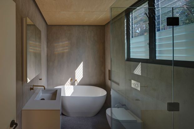 Compact contemporary bathroom with a freestanding white bathtub, wall-mounted vanity and integrated rectangular basin, toilet, frameless glass shower enclosure, and a large dark-framed window. Warm wood-toned wall and ceiling finishes contrast with grey tiled or stone-like surfaces and a textured dark floor. Natural light creates striped reflections across the walls.