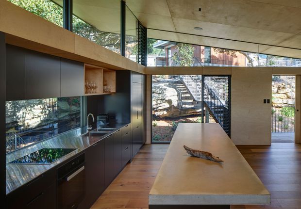 A contemporary open-plan kitchen with a long pale stone island and integrated seating overhang, dark matte cabinetry, reflective dark splashback, timber flooring, and extensive floor-to-ceiling glazing. A warm timber-lined ceiling and large glazed openings connect the kitchen to a rocky landscaped exterior, while a black-framed glazed door and open shelving add architectural detail.