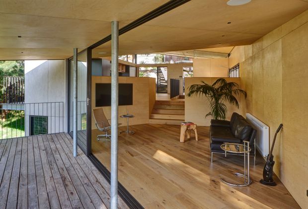A warm, open-plan modern interior viewed from a timber deck, featuring extensive glazing with slender dark frames, plywood-lined walls and ceiling, timber flooring, a compact built-in workspace, a sunken or stepped living area, black leather lounge chairs, and an indoor palm. Slim metal columns and exposed structural elements contribute to the architectural character.