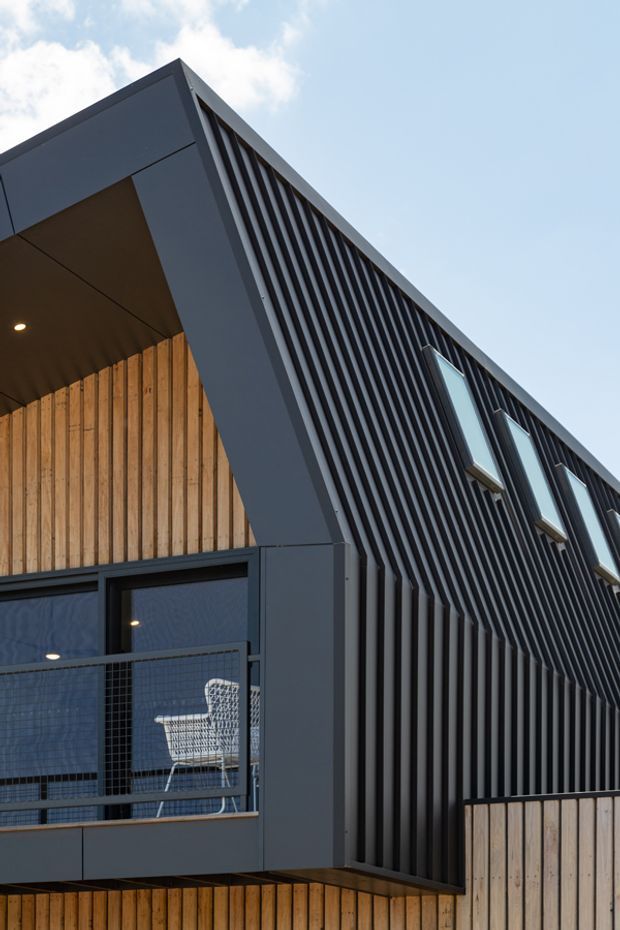 Upward-looking architectural exterior photograph of a contemporary multi-level building. The facade combines dark vertical ribbed cladding and projecting dark window frames with warm vertical timber wall lining and a recessed balcony. A wire-mesh balcony guard and outdoor chair are visible, with a bright blue sky beyond.