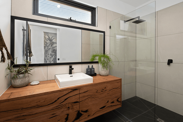 Contemporary bathroom featuring a large timber vanity with distinctive natural grain, a white vessel basin, black tapware and framed mirror, alongside a frameless glass walk-in shower with large light neutral wall tiles and dark floor tiles. Potted plants and restrained accessories add a natural, refined feel.