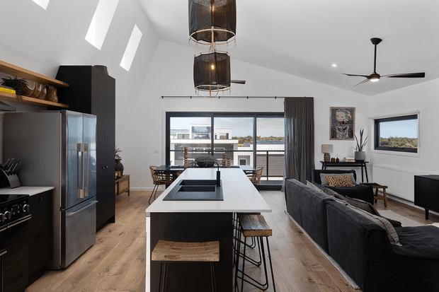 Open-plan contemporary kitchen and living area with a large central island, dark cabinetry, stainless-steel appliances, timber flooring, a charcoal sofa, expansive glazed doors, and a high sloping ceiling with skylights. Black pendant lights and ceiling fans provide strong modern visual accents.