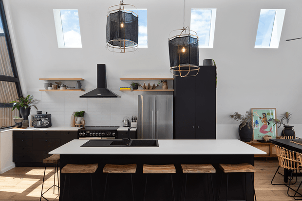 A bright contemporary kitchen with a large white island and dark base cabinetry, integrated cooktop, black rangehood, open timber shelves, stainless-steel appliances, and several pendant lights. Skylights and large windows provide strong natural light, while stools line the island and a small dining area is visible to the right.