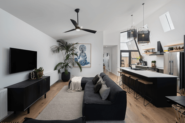 A bright, open-plan contemporary living and kitchen area with white walls, light timber flooring, a dark sectional sofa, black media cabinet, and a large kitchen island with bar seating. The space features black-framed glazing, pendant lights with woven shades, a ceiling fan, indoor plants, and minimal modern furnishings.