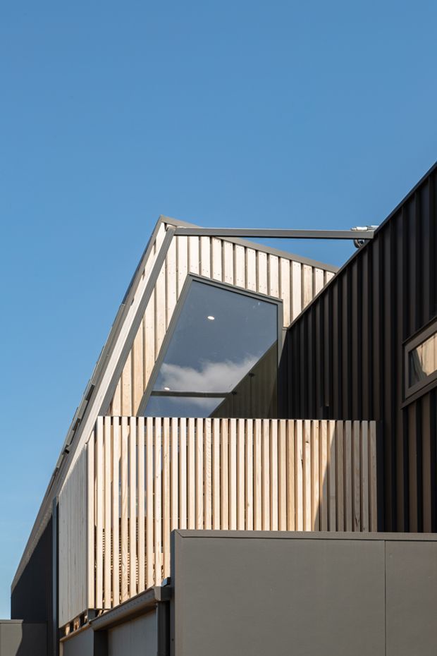 Exterior architectural photograph of a contemporary multi-level building under a clear blue sky. The facade combines light vertical timber screening and cladding with dark ribbed metal cladding, angular pitched roof forms, projecting balconies, and large dark-framed glazing. The composition emphasizes strong geometric lines and contrasting materials.