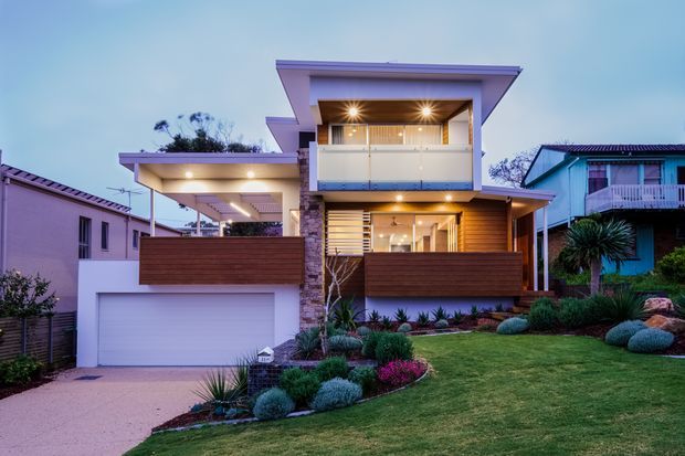 Photograph of a contemporary multi-level suburban residence at dusk, featuring a strong horizontal composition, flat projecting roof forms, warm timber cladding, stone detailing, extensive upper-level glazing with a balcony, an integrated garage, covered outdoor areas, and carefully landscaped front gardens with sculptural planting.