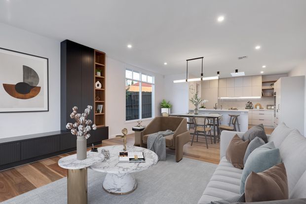 A bright, contemporary open-plan living room flowing into a dining area and kitchen. The room features a large pale sectional sofa, sculptural coffee tables with marble-like tops, neutral upholstered armchairs, timber flooring, a dark vertical-panel media or storage wall, white cabinetry, and linear pendant lighting over the dining island. Large windows provide natural light, while restrained artwork and neutral textiles create a refined, comfortable interior.