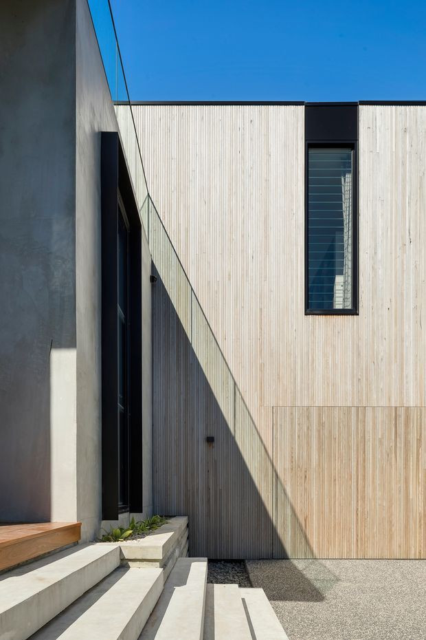 A contemporary residential exterior courtyard or entry passage with pale vertically ribbed timber cladding, smooth grey concrete walls and stepped concrete paving. A tall narrow black-framed window and dark entry door create strong contrast, while sharp sunlight casts diagonal shadows across the facade. Minimal planting and exposed aggregate paving reinforce the clean, modern architectural character.