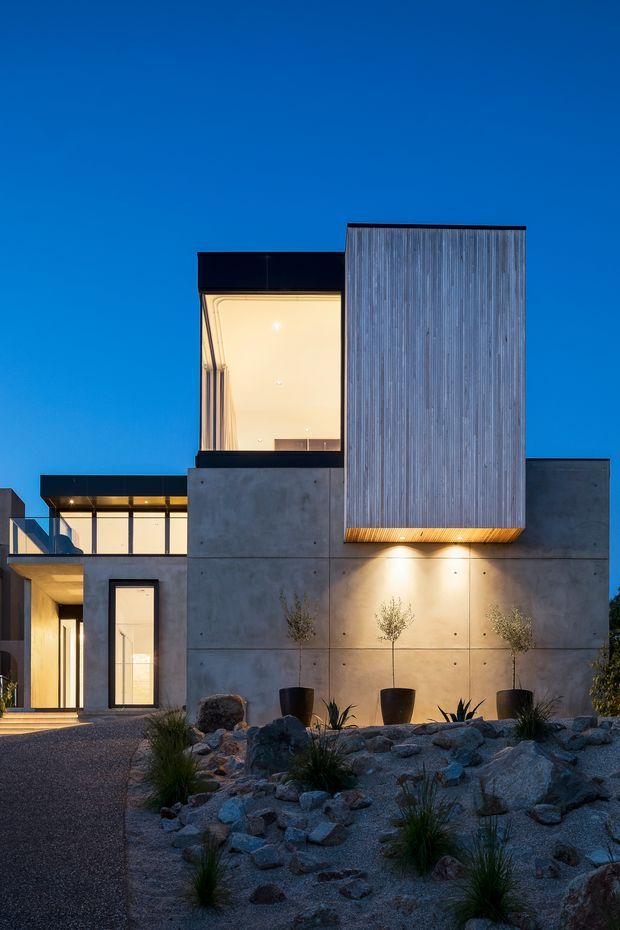 Twilight exterior view of a contemporary multi-level residence with a strong geometric composition. The facade combines smooth exposed concrete panels, dark-framed glazing, and a tall vertically ribbed cladding volume projecting above the main structure. Warm interior and soffit lighting contrasts with the cool blue evening sky. A minimalist xeriscaped front garden uses gravel, rocks, grasses, and potted plants, with a paved approach leading to the entrance.