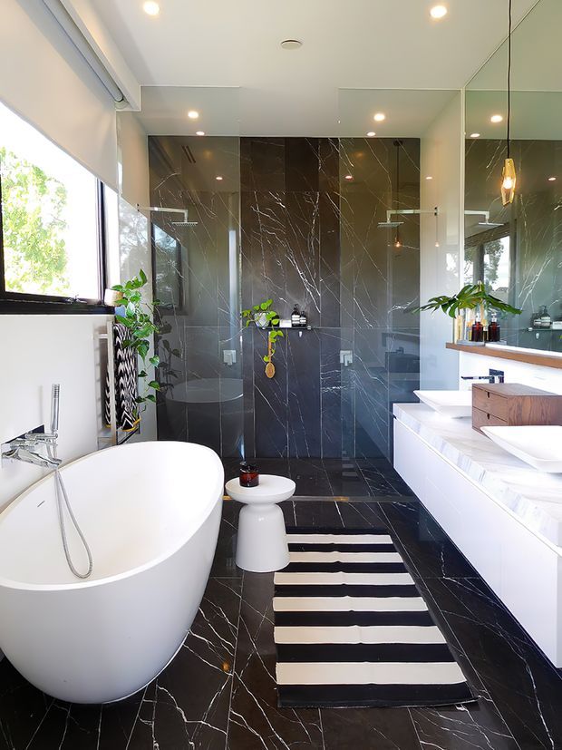 A bright, contemporary bathroom featuring a freestanding white bathtub, wall-mounted white vanity with integrated basins, a glass-enclosed shower, and a wall-hung toilet. Large dark marble-look tiles with prominent white veining cover the shower walls and floor, complemented by white cabinetry, mirrored walls, indoor plants, and a black-and-white striped rug. A large window provides abundant natural light.