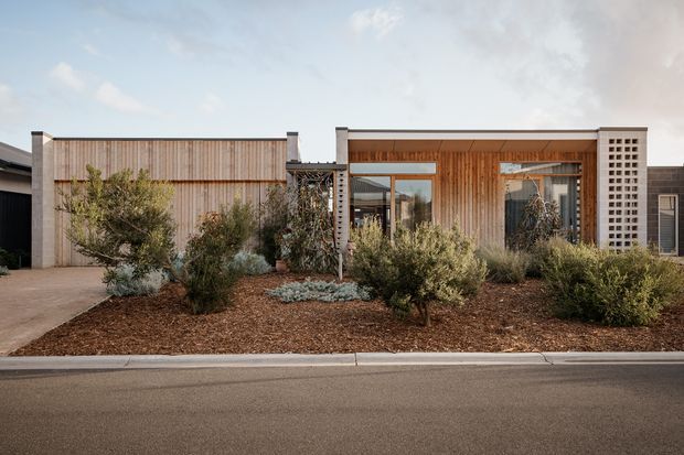 Street-facing view of a low, single-storey contemporary residence with a restrained rectilinear facade. Vertical timber cladding and warm timber screening frame the recessed entry and glazing, while pale rendered or masonry wall sections and perforated breeze-block panels provide contrast. The front garden uses drought-tolerant shrubs, mulch and gravel-like planting beds, with a driveway and kerb visible in the foreground.