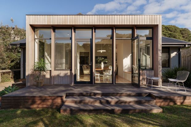 Front exterior view of a compact contemporary home extension with a warm vertical timber-clad facade, extensive floor-to-ceiling glazing, dark framed doors and windows, and a raised timber deck with steps. Outdoor chairs and potted plants sit on the deck, while a bright open-plan interior is visible through the glazing. The building is set in a landscaped suburban garden under a clear sky.
