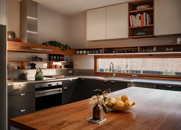 Warm, carefully detailed contemporary kitchen with dark lower cabinetry, pale upper cupboards, timber-framed horizontal window, open shelving, and a large solid timber island. Integrated stainless-steel appliances, a timber display ledge, and curated ceramics and books create a refined, inviting atmosphere.