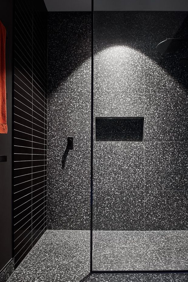 Close-up view of a dark, contemporary walk-in shower. The walls and floor are clad in speckled charcoal terrazzo-like tiles, with a black linear-tile accent wall, recessed wall niche, black shower controls, and a partially visible overhead shower. Dramatic directional lighting highlights the textured tile surface.