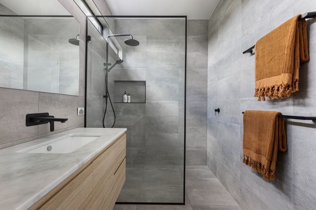 Contemporary bathroom featuring large-format grey stone-look wall and floor tiles, a frameless glass walk-in shower with dual rainfall showerheads, a recessed wall niche, wall-mounted black tapware, and a floating light timber vanity with a pale stone benchtop. Burnt orange towels add warmth and contrast.