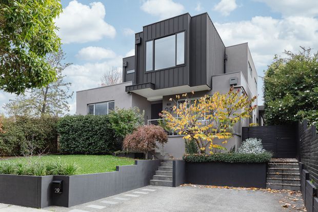Contemporary multi-level urban residence viewed from the street, featuring a dark vertically clad projecting upper volume, pale rendered walls, large windows, stepped concrete entry landscaping, and a dark pedestrian gate and boundary fencing. Mature planting and a small front lawn soften the modern facade.