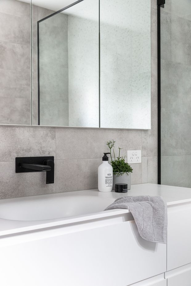 A contemporary bathroom vanity with a white integrated basin and wall-mounted matte-black tapware. Large grey stone-look wall tiles surround the vanity, with a frameless mirrored cabinet above. A white soap dispenser, small potted plant and grey towel add understated styling.
