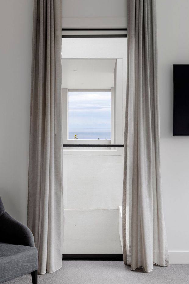 A narrow, bright interior view focused on a tall window opening with layered white architectural framing and a distant ocean view. Full-height textured grey-beige curtains frame the window, with a grey upholstered sofa partially visible at lower left, pale carpet underfoot, and the edge of a wall-mounted black television at right. The space has a clean, restrained contemporary appearance.