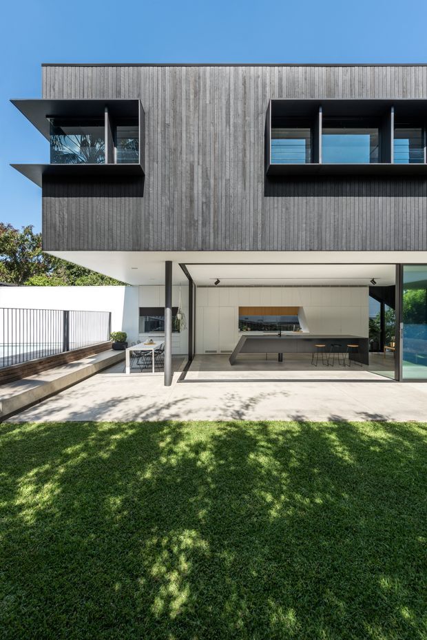 A contemporary two-storey residence viewed from the garden, with a dark vertically clad upper level projecting over a partially open ground-floor kitchen and patio. Large glazed openings, deep black window reveals, a long island bench, concrete outdoor paving, and a manicured lawn create a clean indoor-outdoor connection. The image is photographed in bright daylight against a clear blue sky.