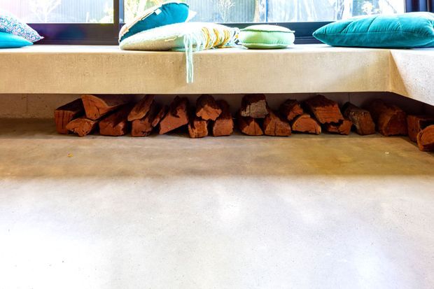 Close-up interior view of a built-in concrete window seat or bench with colourful cushions, featuring an open recessed wood-storage compartment beneath. The surrounding floor and bench surfaces have a smooth, warm-toned concrete finish, with bright daylight entering through the window.