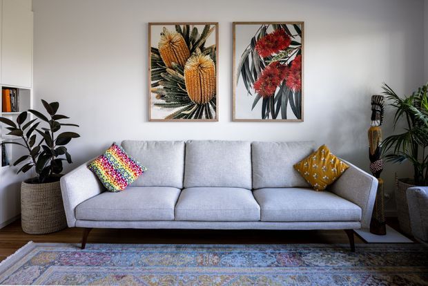 A bright, inviting living room centered on a light grey upholstered sofa with slim dark timber legs. The room features white walls, botanical artwork in timber frames, a patterned area rug, colorful accent cushions, indoor plants, a woven planter, and a tall decorative sculptural object. Built-in shelving with books is visible at left.