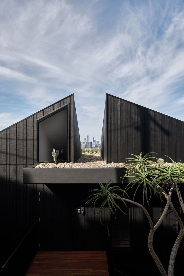 Exterior architectural photograph of a dark, sharply geometric multi-level building. Two tall angular volumes with sloping rooflines frame a narrow rooftop opening containing gravel and sculptural planting, while a deep shadowed entrance or lower-level void sits beneath. Black vertical cladding dominates the facade, with a timber deck at the foreground and a distant urban skyline visible through the central gap.