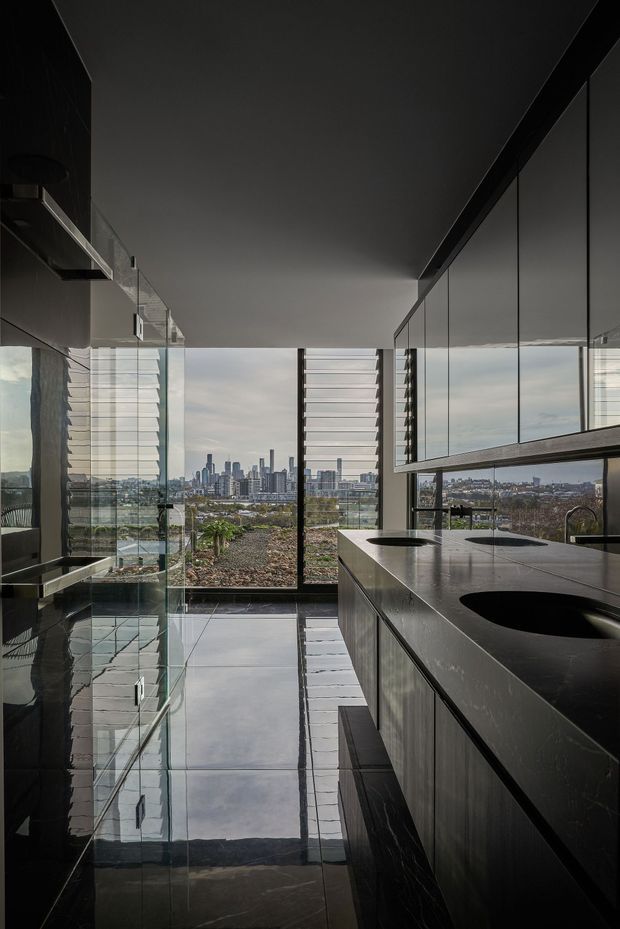A dramatic contemporary kitchen interior with a long dark island, integrated black sink, reflective dark cabinetry, mirrored or glass-panelled walls, and full-height glazing overlooking a dense city skyline. Horizontal louvres filter the daylight, while glossy stone-like surfaces and polished flooring create strong reflections. The space has a sleek, highly minimal and monochromatic character.