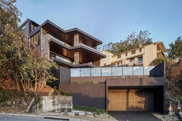 Exterior view of a contemporary multi-level residence on a sloped, landscaped site. The house features projecting balconies and deep roof overhangs, dark textured masonry or stone-clad walls, warm timber soffits, extensive glazing, and a recessed garage with a timber door. A neighbouring multi-storey dwelling is visible behind the property.
