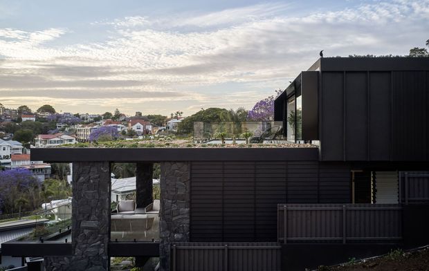 Exterior view of a contemporary multi-level home on a sloped suburban site, featuring dark horizontal cladding, a flat roof, large glazed openings, and rough stone structural columns. A covered outdoor terrace is visible beneath the upper volume, with neighbouring homes and landscaped gardens in the background.