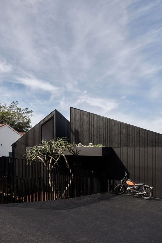 Exterior architectural photograph of a contemporary dark-clad building with sharply angled, folded roof and wall planes. Vertical ribbed cladding forms a strong geometric facade, with a recessed covered entry or parking area behind a dark slatted fence. A planted terrace edge and mature tree soften the composition, while a motorcycle sits in the foreground driveway. Bright sky and strong sunlight create pronounced shadows.